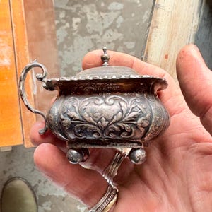 May include: An antique silver sugar bowl with a lid and handle. The bowl has ornate floral and leaf patterns and sits on three small ball feet. The handle is curved, and the lid has a small finial. The bowl is held in a person's hand.