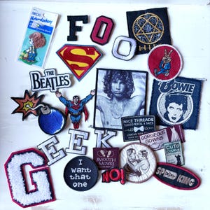 May include: A selection of embroidered patches with diverse designs and text. Includes a Superman logo, a Beatles logo, a portrait of Jim Morrison, and the word "BOWIE". Other patches feature the words "FOO", "GEEK", and "SPEED KING".