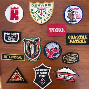 May include: A collection of thirteen embroidered fabric patches in various shapes and colours. The patches feature diverse logos, text including 'Toro', 'Coastal Patrol', 'Intermodal', 'Masterton', and 'Westpoint Cleaning Staff', and graphic illustrations.