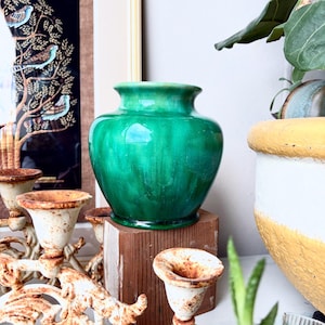 May include: A vibrant, emerald green vase with a rounded form and a flared rim. The vase rests on a small, square wooden block. Other decor includes a white and gold planter and a vintage candelabra.
