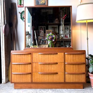 May include: A wooden chest of drawers featuring a rectangular mirror attached to the top. The unit has three vertical sections with three drawers in each, totalling nine drawers. Each drawer is fitted with a horizontal, curved wooden handle.