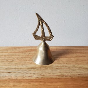 May include: A brass bell with a sailboat design. The bell is on a wooden surface. The sailboat has a detailed design, and the bell has a classic shape. The bell is made of brass.