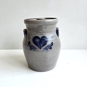 May include: A gray stoneware crock with a lid, featuring two side handles and a dark blue hand-painted heart design. The front displays a stamped logo reading Iowa Pottery Works, Cambridge, WI, and the year 1993 in dark blue ink.