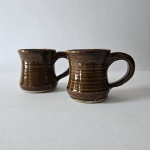 May include: Two handmade ceramic mugs with a glossy dark brown glaze. Each mug features a cylindrical body with subtle horizontal ridges, a wide flared rim, and a sturdy curved handle. The base of each mug is unglazed, showing a light beige clay.