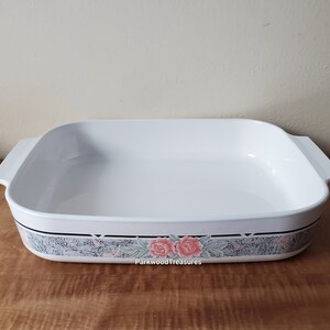 May include: White ceramic baking dish with a floral design. The design features two pink roses with green leaves on a gray and white background. The dish has two handles on the sides.
