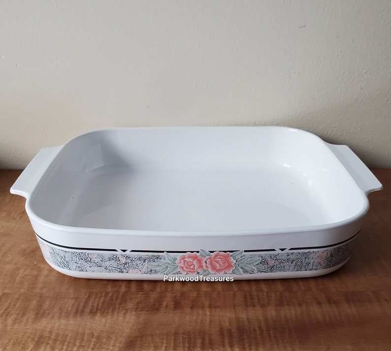 May include: White ceramic baking dish with a floral design. The design features two pink roses with green leaves on a gray and white background. The dish has two handles on the sides.