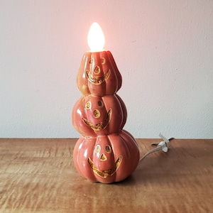 May include: A ceramic lamp shaped like a stack of three orange jack-o'-lanterns with carved faces. The top pumpkin features a glowing light bulb protruding from the top. The lamp has a white electrical cord extending from the base to the right side.
