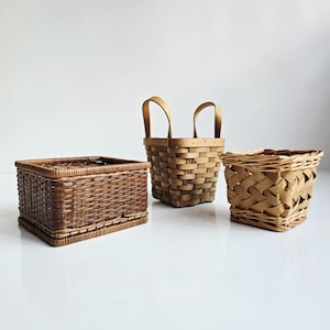 May include: Three woven baskets of varying sizes and styles. The basket on the left is rectangular with a dark brown finish. The center basket has a square shape with two handles. The basket on the right has a woven pattern.