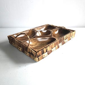 May include: A rectangular wooden serving tray with five compartments. The tray has a woven border and rope handles. The wooden compartments are a medium brown color, and the woven border is a mix of light and dark brown.