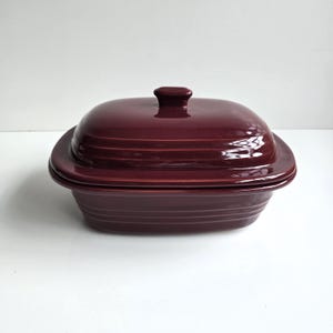 May include: A deep, rectangular, dark maroon ceramic casserole dish with a matching lid. The dish features a glossy finish and horizontal ribbed detailing along the sides of both the base and the lid, which includes a central rectangular handle.