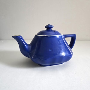 May include: A cobalt blue ceramic teapot with a glossy finish, featuring a faceted, multi-sided body, a curved spout, a sturdy handle, and a removable lid with a small knob handle. The base and lid rim are accented with a thin white line.