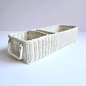May include: A rectangular, white wicker basket with two interior compartments and a handle on each end. The basket is painted white and appears to be made of woven wicker. The basket is designed for storage and organization.
