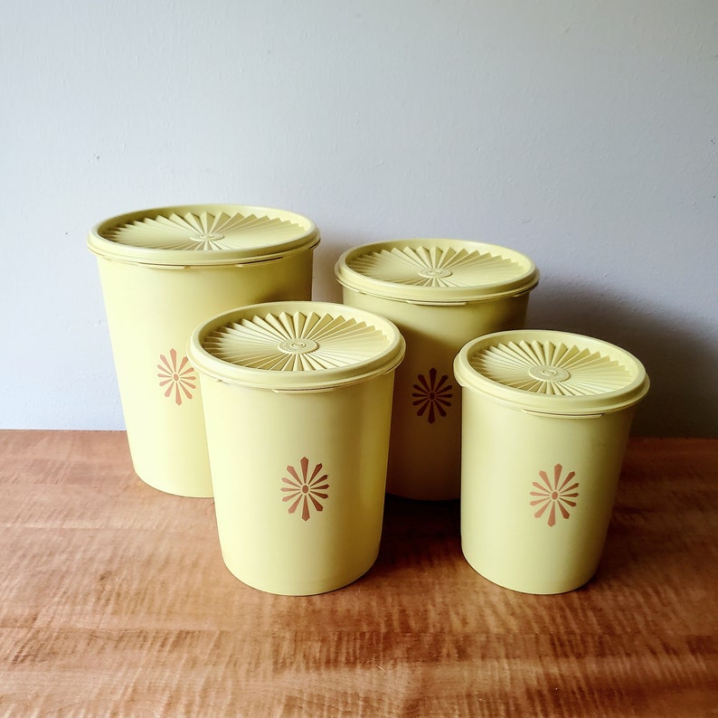 K&ouml;nnte beinhalten: Ein Set aus vier gelben Vintage-Kunststoffbeh&auml;ltern mit passenden Deckeln. Jeder Beh&auml;lter hat ein braunes Blumendesign und einen geriffelten Deckel. Die Beh&auml;lter sind in verschiedenen Gr&ouml;&szlig;en auf einer Holzoberfl&auml;che angeordnet.
