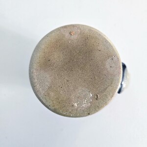 Op de afbeelding: Een steengoed mok met een gespikkelde, lichtbruine buitenkant en een donkerblauwe handgreep. De bodem van de mok is zichtbaar en toont een getextureerd oppervlak met kleine onvolkomenheden en het nummer "9" gestempeld.