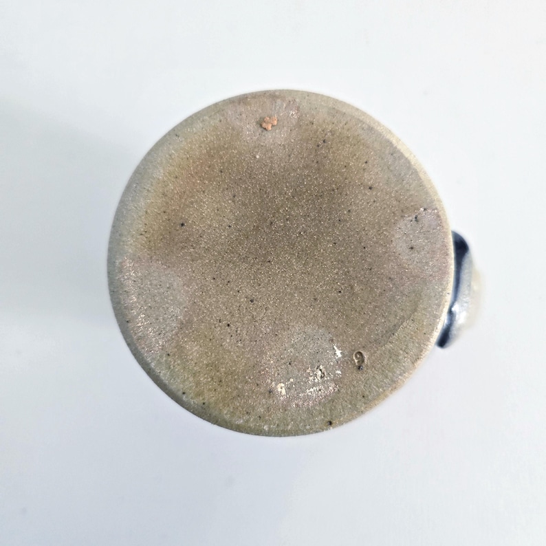 Op de afbeelding: Een steengoed mok met een gespikkelde, lichtbruine buitenkant en een donkerblauwe handgreep. De bodem van de mok is zichtbaar en toont een getextureerd oppervlak met kleine onvolkomenheden en het nummer "9" gestempeld.