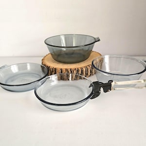 May include: A set of four vintage glass bowls in shades of blue and gray. The bowls vary in size and shape, with some featuring handles. A black and clear handle is also included. The bowls are arranged on a white surface and a wooden slice.