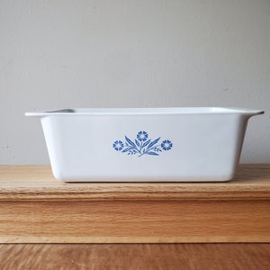 Puede incluir: Molde para pan de cer&aacute;mica blanca con un dise&ntilde;o floral azul. El dise&ntilde;o presenta tres flores azules con tallos y hojas verdes.