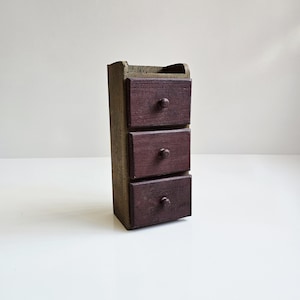 May include: A small, wooden cabinet with three drawers. The drawers are a deep burgundy color with round knobs. The cabinet's frame is a muted green-brown. The piece is likely a miniature storage unit or decorative item.