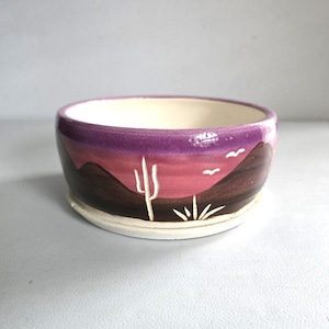 May include: A ceramic bowl with a desert landscape design. The bowl features a purple rim, a sunset scene with mountains, a cactus, and birds. The interior is off-white, and the base is a lighter shade of white.
