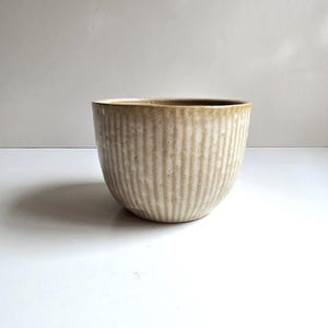 May include: A ceramic bowl featuring a vertical ribbed texture and a reactive glaze finish in shades of beige, cream, and light brown. The bowl has a wide, open top and a rounded base, showcasing a subtle gradient of earthy tones across its surface.