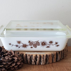 May include: White glass baking dish with a brown pine cone design and a matching lid. The dish is sitting on a wooden surface.