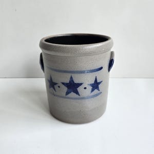 May include: A cylindrical gray stoneware crock featuring a dark blue painted band with three five-pointed stars and small dots between them. The vessel has a thick rim, a smooth exterior finish, and two small side handles with dark blue accents.