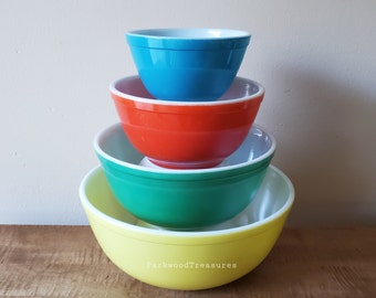 Puede incluir: Un juego de cuatro tazones para mezclar anidados en amarillo, verde, naranja y azul. Los tazones son de vidrio y tienen un borde blanco.