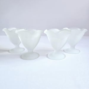 May include: Four frosted glass dessert cups. Each cup has a tulip-shaped bowl with a ruffled rim, set on a pedestal base. The cups are a soft, opaque white, and are arranged on a white surface.