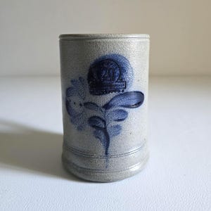 May include: A cylindrical stoneware mug with a light gray base color and blue floral design. The mug features a blue emblem with the number 20. The mug has a textured surface and a slightly flared base.