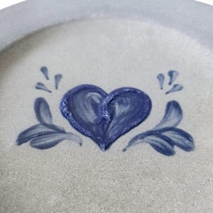 K&ouml;nnte beinhalten: Ein Keramikteller mit hellgrauem Hintergrund und blauem Rand. Der Teller zeigt ein handgemaltes blaues Herzdesign in der Mitte, mit stilisierten blauen floralen Akzenten auf beiden Seiten. Der Teller hat eine rustikale, handgemachte &Auml;sthetik.