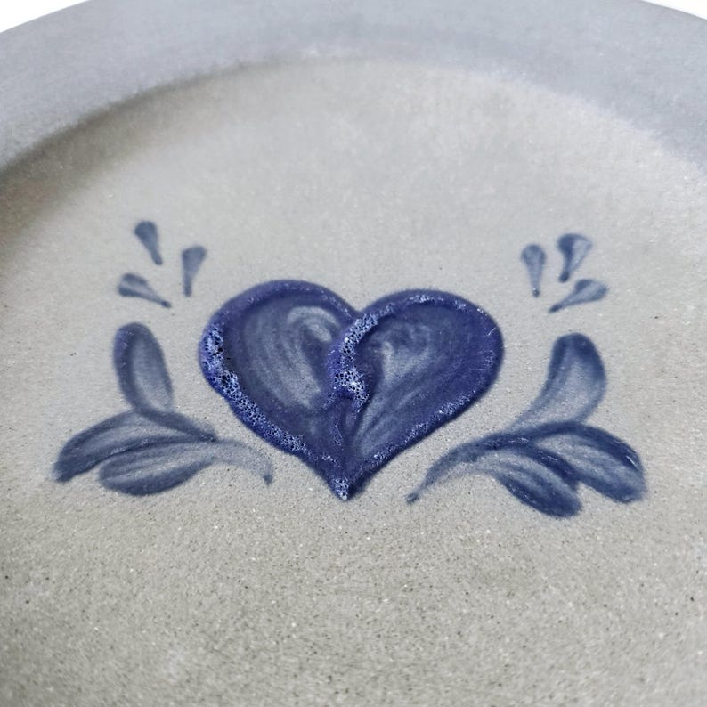 K&ouml;nnte beinhalten: Ein Keramikteller mit hellgrauem Hintergrund und blauem Rand. Der Teller zeigt ein handgemaltes blaues Herzdesign in der Mitte, mit stilisierten blauen floralen Akzenten auf beiden Seiten. Der Teller hat eine rustikale, handgemachte &Auml;sthetik.