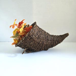 May include: A dark brown woven wicker cornucopia basket filled with artificial autumn foliage in shades of orange, yellow, and brown. The horn-shaped basket features a textured, natural twig construction with a wide opening and a tapered end.