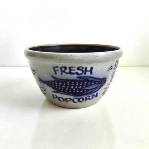 May include: A round ceramic bowl with a speckled light gray exterior and a dark brown interior. The front features a blue graphic of an ear of corn with the text FRESH POPCORN printed in blue lettering above and below the corn illustration.