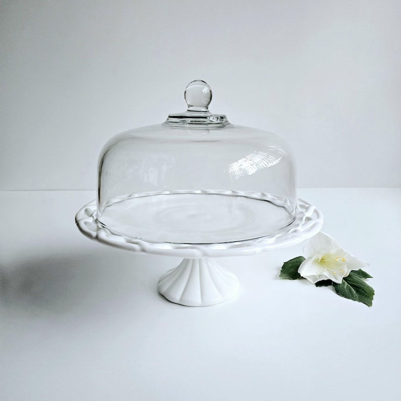 May include: White cake stand with a glass dome. The stand has a fluted base and a scalloped edge. The dome has a knob on top.