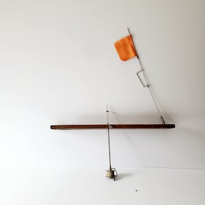 May include: An ice fishing tip-up with a wooden base, metal frame, and an orange flag. The reel is at the bottom, and the flag is angled upwards. The tip-up is designed to signal when a fish bites.