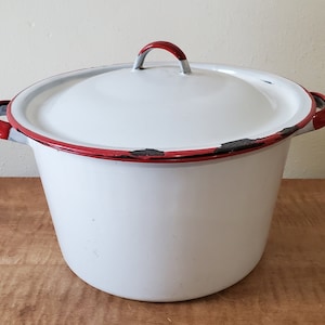 May include: White enamelware pot with a lid. The pot has a red rim and handles, and a red handle on the lid. The enamel is chipped in places, giving it a vintage look. The pot is sitting on a wooden surface.
