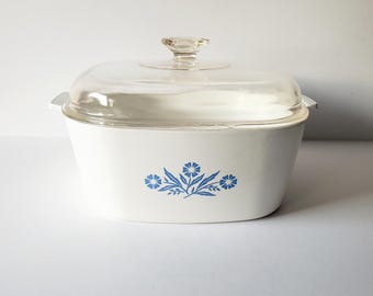 May include: White casserole dish with a glass lid and a blue floral design. The design features three blue flowers with green leaves.