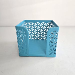 May include: A square, light blue metal desk organizer with a decorative cut-out pattern. The organizer has a solid base and sides with a floral and dot design. The open design allows for easy access to contents.