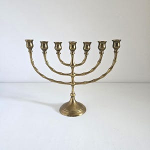 May include: A brass menorah with seven candle holders. The base is round, and the arms curve upwards, each holding a candle cup. The design has a bamboo-like texture. The menorah is a symbol of the Jewish faith.