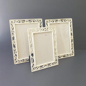 May include: Three off-white picture frames with ornate, scrolling designs. The frames are rectangular, with varying sizes, and are arranged on a dark gray surface. The frames have a vintage or antique aesthetic.