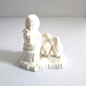 Puede incluir: Figura de cer&aacute;mica blanca que representa un mu&ntilde;eco de nieve sosteniendo un regalo, con dos ping&uuml;inos cerca. El mu&ntilde;eco de nieve est&aacute; sobre una base cubierta de nieve. La figura tiene un aspecto texturizado similar a la nieve.