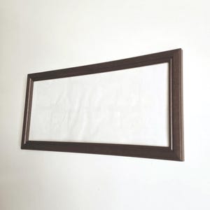 May include: A rectangular brown picture frame with a white interior, mounted on a white wall. The frame has a simple, classic design, suitable for displaying artwork or photographs. The frame is made of wood.