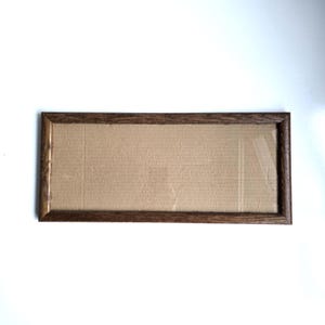 May include: A rectangular picture frame with a dark brown wood grain finish. The frame features mitered corners and contains a plain, light brown cardboard insert that fills the entire interior space, providing a flat and neutral surface.