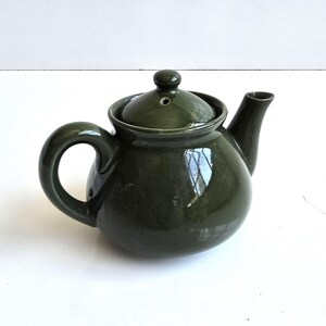 May include: A dark olive green ceramic teapot with a glossy finish, featuring a rounded body, a curved handle, a short spout, and a matching lid with a small steam hole at the top. The teapot has a smooth, reflective surface throughout.