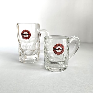 May include: Two clear glass A&W Root Beer mugs. The mugs have a handle and a diamond pattern on the sides. Each mug has a red, white, and black A&W Root Beer logo. The mugs are on a white surface.
