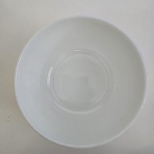 May include: A plain white ceramic bowl viewed from directly above. The bowl features a smooth, glossy finish with a circular base indentation in the center and a simple, rounded rim. The design is entirely minimalist, featuring no patterns or colors.