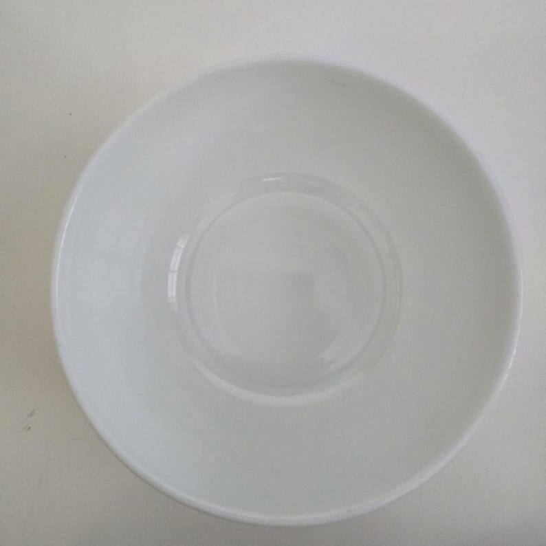 May include: A plain white ceramic bowl viewed from directly above. The bowl features a smooth, glossy finish with a circular base indentation in the center and a simple, rounded rim. The design is entirely minimalist, featuring no patterns or colors.