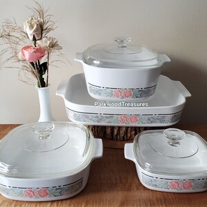 May include: Set of four white casserole dishes with glass lids. The dishes feature a floral pattern with pink roses and green leaves on a gray background. The dishes are arranged in a pyramid shape with the largest dish at the bottom and the smallest dish at the top.