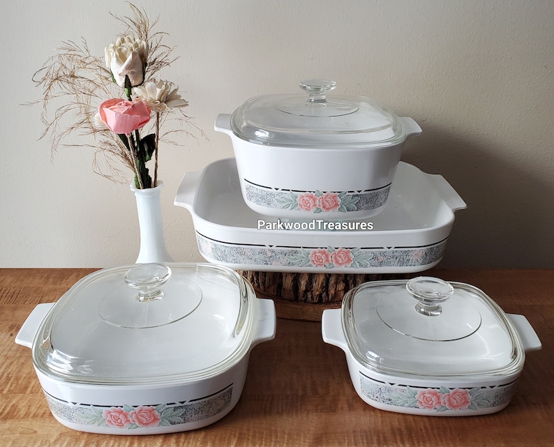 May include: Set of four white casserole dishes with glass lids. The dishes feature a floral pattern with pink roses and green leaves on a gray background. The dishes are arranged in a pyramid shape with the largest dish at the bottom and the smallest dish at the top.