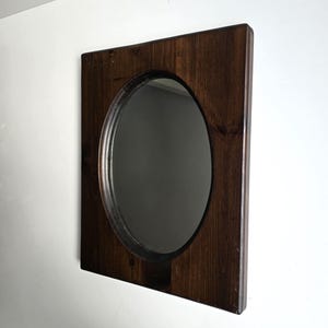 May include: A square wall mirror featuring a dark brown stained wooden frame with a visible wood grain texture. The mirror glass is oval-shaped and set into the center of the square wooden panel, creating a contrast between the rounded reflective surface and the straight edges of the frame.
